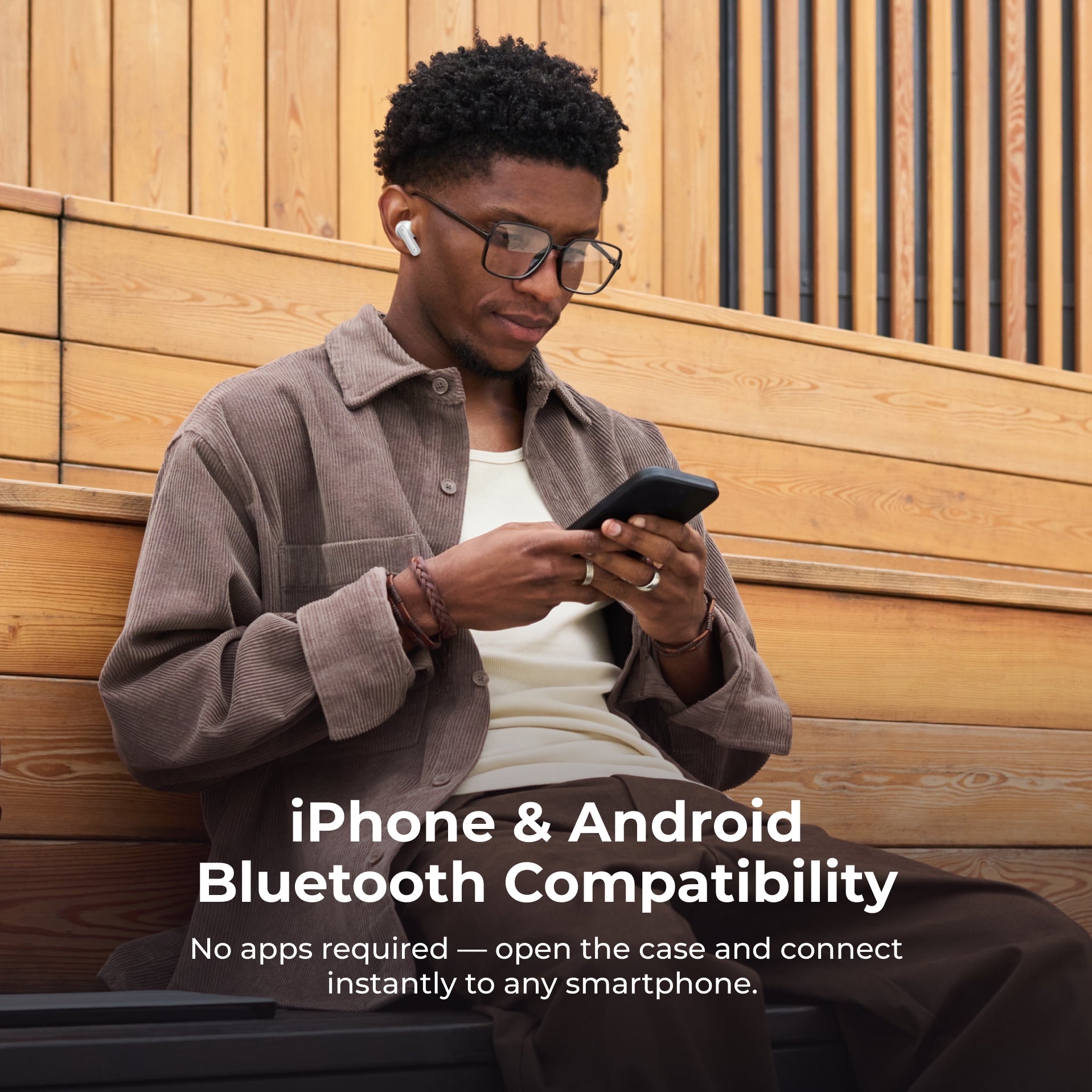 Man using a smartphone with Bluetooth headphones, sitting against a wooden wall.